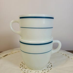 May include: Two stacked white milk glass mugs featuring a smooth, glossy finish and two thin, horizontal dark blue stripes encircling the upper portion of each cup. Each mug has a C-shaped handle extending from the side of the rounded body.