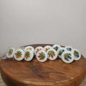 May include: A collection of round white ceramic cabinet knobs featuring various botanical illustrations. Each knob displays a unique floral or plant motif in shades of orange, green, and blue. Some knobs are packaged in clear plastic.