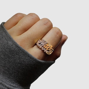 May include: A gold-colored ring with the name "Macey" in a cursive font. The ring is worn on a finger, and the name is embellished with small, sparkling details. The ring is a personalized piece of jewelry.