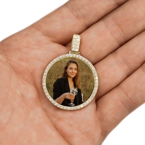 May include: A gold pendant with a round photo frame encrusted with clear cubic zirconia stones. The photo inside the frame shows a woman holding a glass of wine.