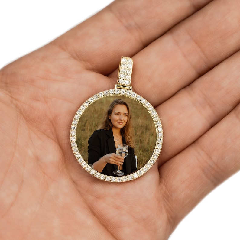 May include: A gold pendant with a round photo frame encrusted with clear cubic zirconia stones. The photo inside the frame shows a woman holding a glass of wine.