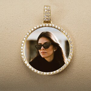 May include: A gold pendant with a round photo frame encrusted with small diamonds. The frame contains a photo of a woman wearing sunglasses.