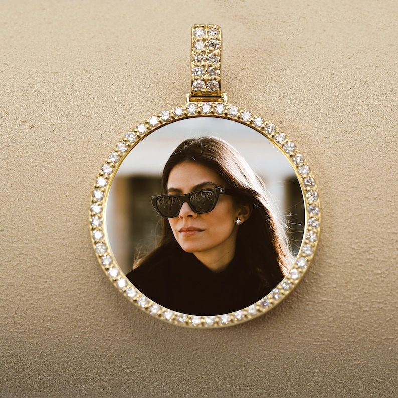 May include: A gold pendant with a round photo frame encrusted with small diamonds. The frame contains a photo of a woman wearing sunglasses.