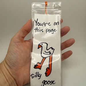 May include: A rectangular white paper bookmark encased in a clear plastic sleeve with an orange string at the top. The bookmark features a hand-drawn illustration of a white goose with orange legs and beak, with the text "You're on this page" and "silly goose" written in black ink.