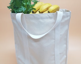 May include: A white canvas tote bag with two long handles. The bag is filled with bananas and celery.