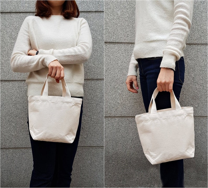 May include: Two people are holding a plain white canvas tote bag with short handles. The bag is made of natural cotton fabric and has a simple design.