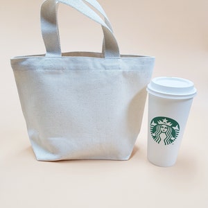 May include: A white canvas tote bag with two handles and a white coffee cup with a green Starbucks logo.