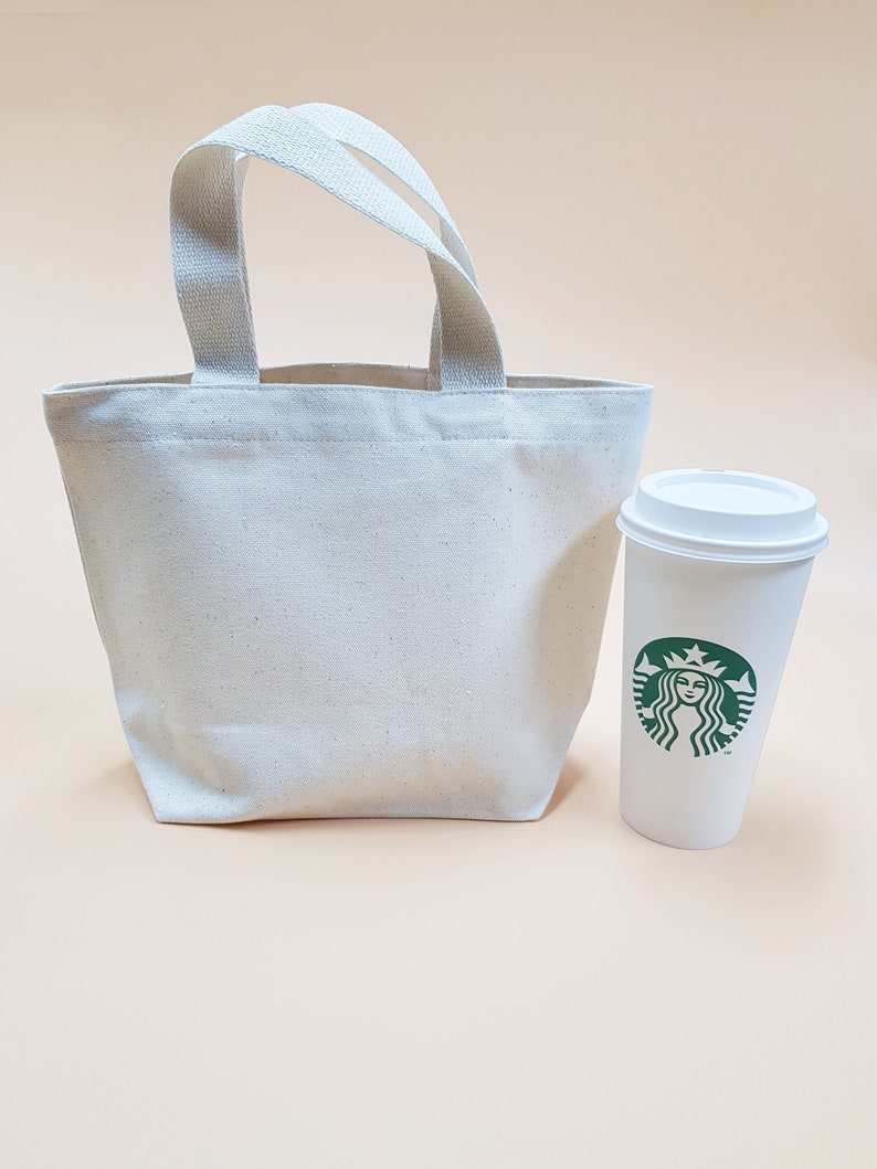 May include: A white canvas tote bag with two handles and a white coffee cup with a green Starbucks logo.