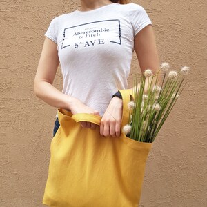 Pode incluir: Uma mulher está segurando uma bolsa tote amarela mostarda com um buquê de flores secas. A bolsa tote é feita de um tecido natural. A mulher está vestindo uma camiseta branca com o texto "Abercrombie & Fitch 5th AVE" impresso nela.