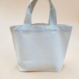 May include: A plain white canvas tote bag with two handles. The bag is small and would be suitable for carrying small items.