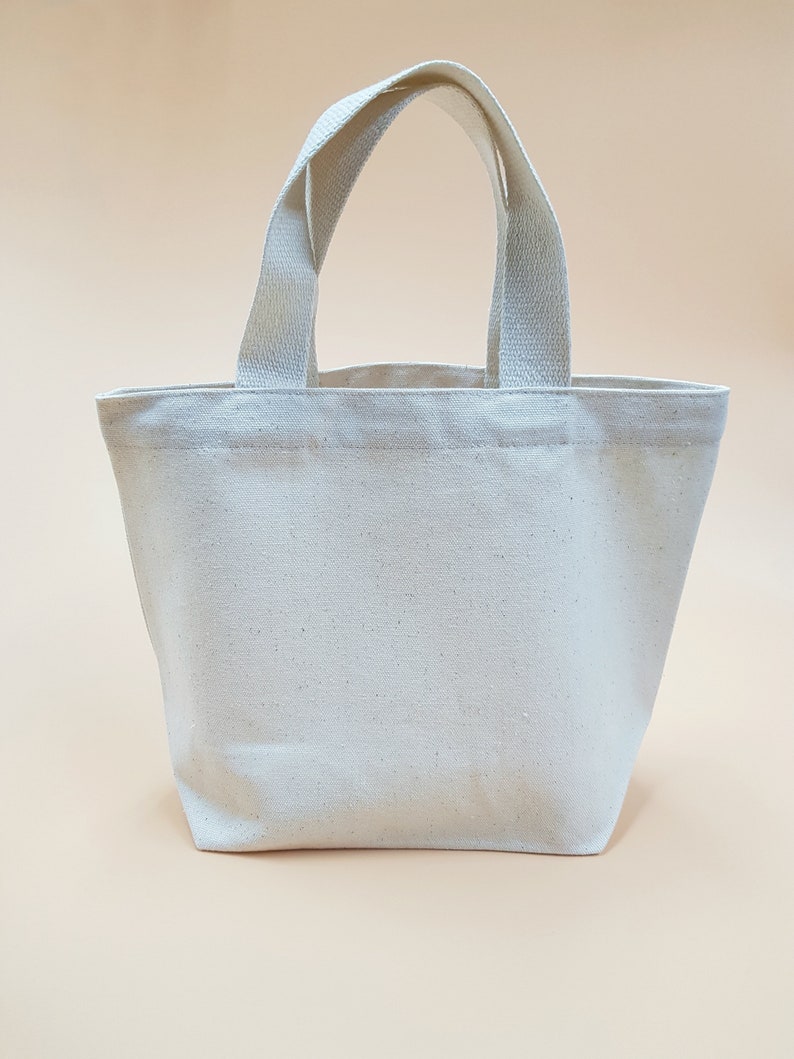 May include: A plain white canvas tote bag with two handles. The bag is small and would be suitable for carrying small items.