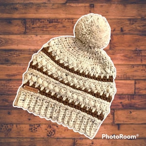May include: A crocheted beanie hat in a brown and white colour scheme. The hat has a pom pom on top and a brown leather tag that says "Fleur de Nivo".