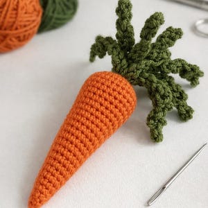 Op de afbeelding: Een handgemaakte gehaakte wortel met groene bladeren. De wortel is oranje en de bladeren zijn groen. Twee bollen wol, één oranje en één groen, staan op de achtergrond. Een naald en een schaar zijn ook zichtbaar.
