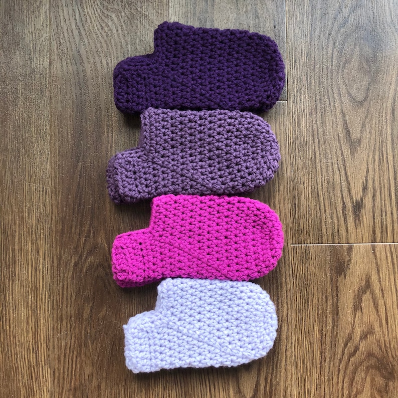 May include: Four crocheted slipper socks in different shades of purple, pink, and white. The socks are arranged vertically, with the darkest shade at the top and the lightest shade at the bottom.