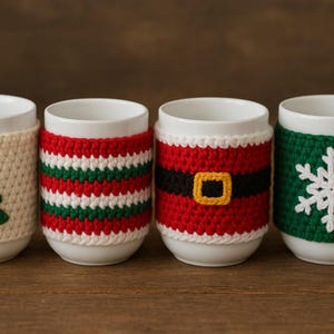 Puede incluir: Cuatro tazas de cerámica blanca con fundas de ganchillo. Las fundas presentan diseños de árbol de Navidad, rayas, cinturón de Papá Noel y copo de nieve. Las tazas están sobre una superficie de madera.