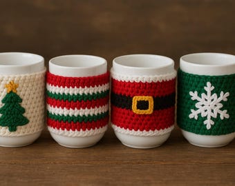 Może przedstawiać: Cztery białe ceramiczne kubki z szydełkowymi osłonami. Osłony mają wzory choinki, paski, paska Świętego Mikołaja i płatka śniegu. Kubki stoją na drewnianej powierzchni.