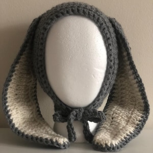 May include: A grey and white crocheted bunny hat with long, floppy ears. The hat is made of soft yarn and has a bow under the chin. The inside of the ears is white, and the edges are grey.