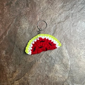 May include: A crocheted watermelon slice keychain. The red fruit has black seed details and a white rind, with a bright yellow-green outer edge. The keychain is attached to a silver metal ring.