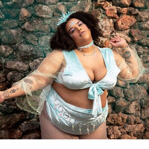 May include: A light blue velvet two-piece outfit with a tie-front top and high-waisted bottoms. The outfit is adorned with a silver chain belt and a matching choker necklace. The model is wearing a crown and has face gems.