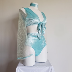 May include: A light blue velvet two-piece outfit on a mannequin. The set includes a tie-front top, high-waisted bottoms, and a choker. One sleeve is made of a sheer, glittery fabric. The outfit is displayed on a white mannequin against a white background.