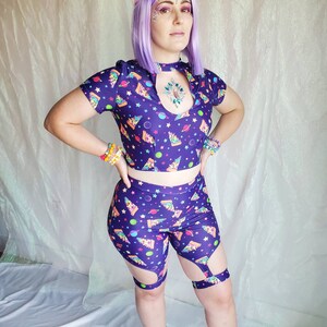 Puede incluir: Un conjunto de top corto y pantalones cortos de color morado y negro con estampado de espacio y pizza. El top tiene un escote de ojo de cerradura y los pantalones cortos tienen un corte de muslo alto.