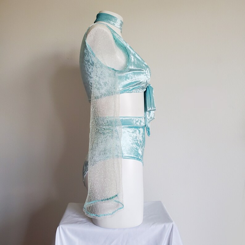 May include: A light blue velvet two-piece outfit on a white mannequin. The set includes a halter top with a tie closure and high-waisted shorts. A sheer, glittery sleeve extends from the top. The outfit is displayed on a white mannequin.
