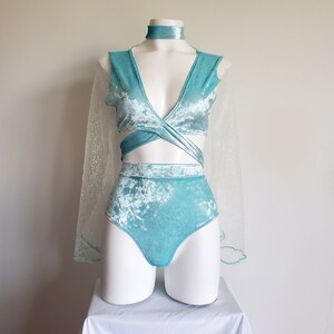 May include: A light blue velvet dance costume set on a mannequin. The set includes a wrap top, high-waisted shorts, a choker, and a sheer, glittery cape. The costume is displayed on a white surface.
