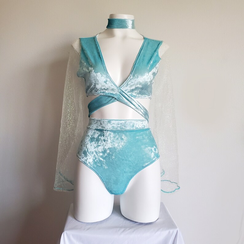 May include: A light blue velvet dance costume set on a mannequin. The set includes a wrap top, high-waisted shorts, a choker, and a sheer, glittery cape. The costume is displayed on a white surface.