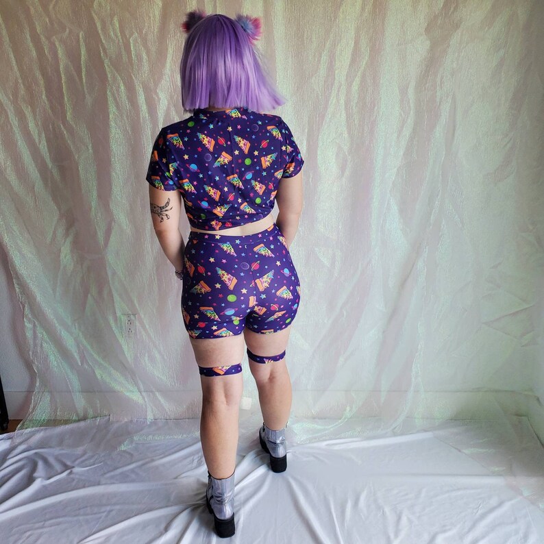 Puede incluir: Un top corto morado y pantalones cortos a juego con un estampado de pizza espacial. El top tiene mangas cortas y un lazo en la parte delantera. Los pantalones cortos tienen cintura alta y est&aacute;n hechos de una tela el&aacute;stica.