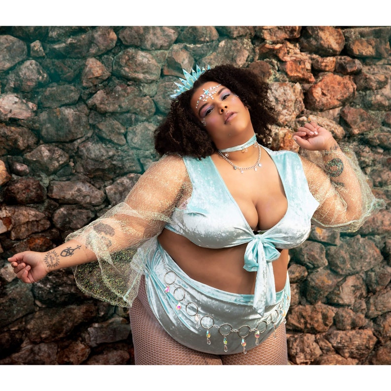 May include: A light blue velvet two-piece outfit with a tie-front top and high-waisted bottoms. The outfit is adorned with a silver chain belt and a matching choker necklace. The model is wearing a crown and has face gems.