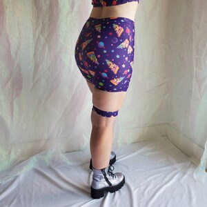 Puede incluir: Pantalones cortos de color morado con un estampado de tem&aacute;tica espacial que incluye rebanadas de pizza, planetas y estrellas. Los pantalones cortos tienen una cintura el&aacute;stica ancha y se llevan con botas de plataforma plateadas.