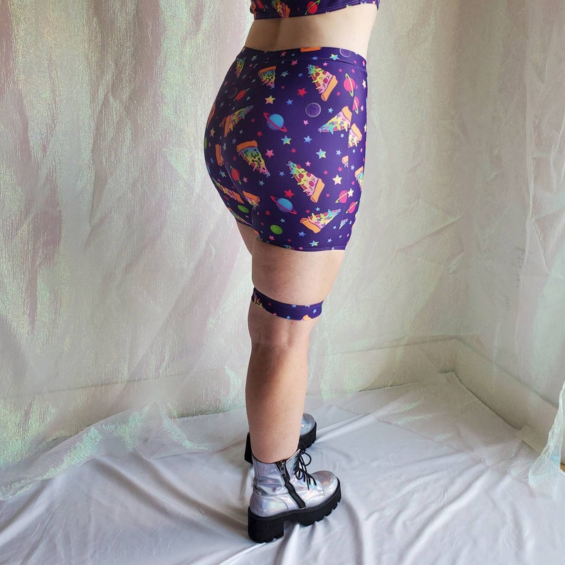 Puede incluir: Pantalones cortos de color morado con un estampado de tem&aacute;tica espacial que incluye rebanadas de pizza, planetas y estrellas. Los pantalones cortos tienen una cintura el&aacute;stica ancha y se llevan con botas de plataforma plateadas.