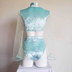 May include: A light blue velvet two-piece outfit on a mannequin. The set includes a crop top, high-waisted shorts, and a matching choker. Sheer, glittery fabric extends from the sleeves. The outfit is displayed on a white mannequin against a plain background.
