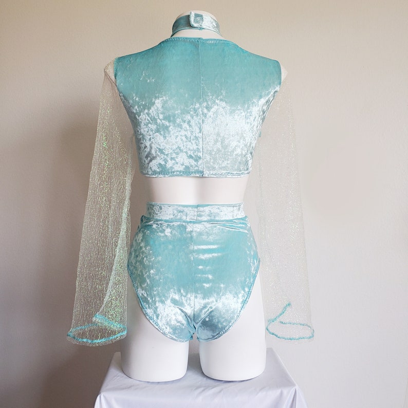 May include: A light blue velvet two-piece outfit on a mannequin. The set includes a crop top, high-waisted shorts, and a matching choker. Sheer, glittery fabric extends from the sleeves. The outfit is displayed on a white mannequin against a plain background.