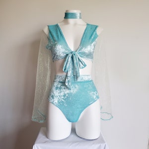 May include: A turquoise velvet two-piece outfit displayed on a mannequin. The set includes a tie-front top, high-waisted bottoms, and a choker. Sheer, glittery sleeves are attached to the top. The outfit is displayed on a white mannequin against a white background.