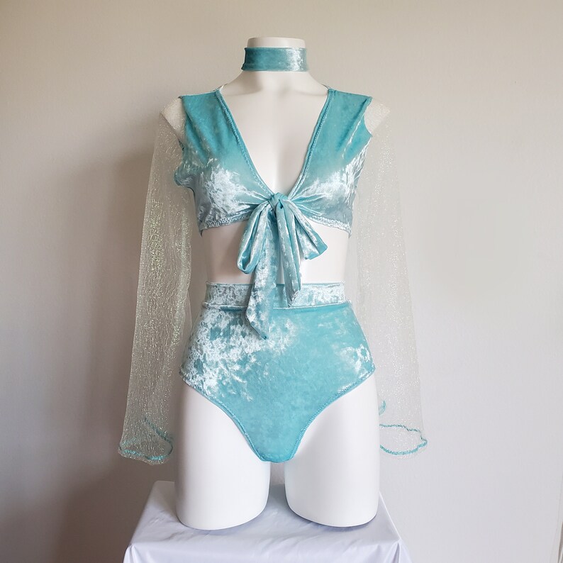 May include: A turquoise velvet two-piece outfit displayed on a mannequin. The set includes a tie-front top, high-waisted bottoms, and a choker. Sheer, glittery sleeves are attached to the top. The outfit is displayed on a white mannequin against a white background.