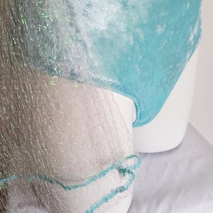 May include: A light blue bodysuit with a delicate, textured fabric. The bodysuit is partially covered by a sheer, silver-white fabric with iridescent threads. The edges of the sheer fabric are trimmed with a turquoise thread.