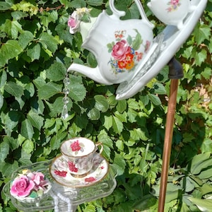 May include: A white porcelain teapot with floral designs pours into a white teacup with a floral design on a glass tray. A white porcelain teacup with floral designs sits on a glass tray with a pearl necklace. The teapot and teacup are attached to a metal pole.