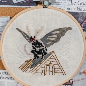 May include: A circular wooden embroidery hoop containing a piece of off-white fabric. The fabric features a hand-stitched design of a dark, winged creature with bright red eyes perched atop a brown-outlined house roof with a crescent moon behind.