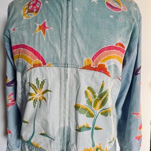 May include: Light blue zip-up jacket with a colorful, whimsical design. The jacket features illustrations of planets, stars, rainbows, and palm trees. The design is printed on the front and sleeves of the jacket. The jacket has a stand-up collar and two snap closures at the bottom.