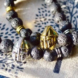 Puede incluir: Dos pulseras de cuentas de piedra de lava negra con detalles dorados y plateados. Cada pulsera presenta un dije decorativo de casco. Las pulseras descansan sobre una pequeña bolsa de tela estampada.
