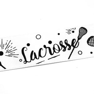 以下が含まれることがあります： ラクロススティック、ボール、そして筆記体で書かれた「Lacrosse」という文字が黒色で繰り返しプリントされた白いリボン。デザインには小さな円形のアクセントと、ボールの周りに放射状の線が含まれており、連続したパターンを形成しています。