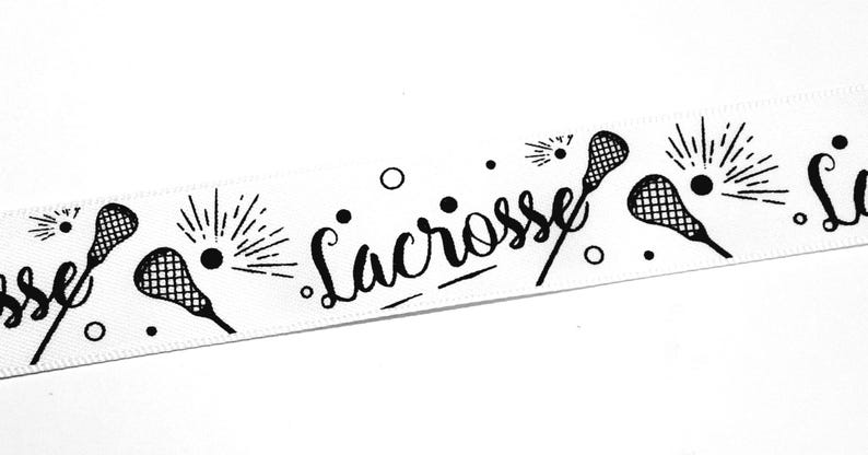 以下が含まれることがあります： ラクロススティック、ボール、そして筆記体で書かれた「Lacrosse」という文字が黒色で繰り返しプリントされた白いリボン。デザインには小さな円形のアクセントと、ボールの周りに放射状の線が含まれており、連続したパターンを形成しています。