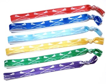 May include: A set of six elastic hair ties in red, blue, light blue, yellow, green, and purple. Each hair tie features a repeating white graphic pattern of two crossed lacrosse sticks and the word lacrosse printed along the fabric surface.