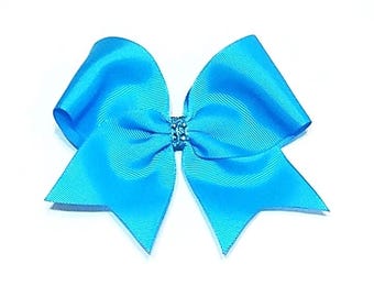 May include: A bright turquoise hair bow made of ribbed fabric with a central knot detail featuring small, clear, sparkling rhinestones. The bow has two large, symmetrical loops and two pointed tails extending downwards from the centre knot.