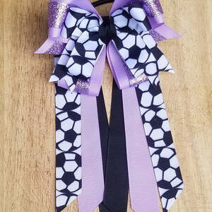 May include: Purple and black hair bow with a soccer ball pattern. The bow has a glitter accent and is attached to a black elastic.