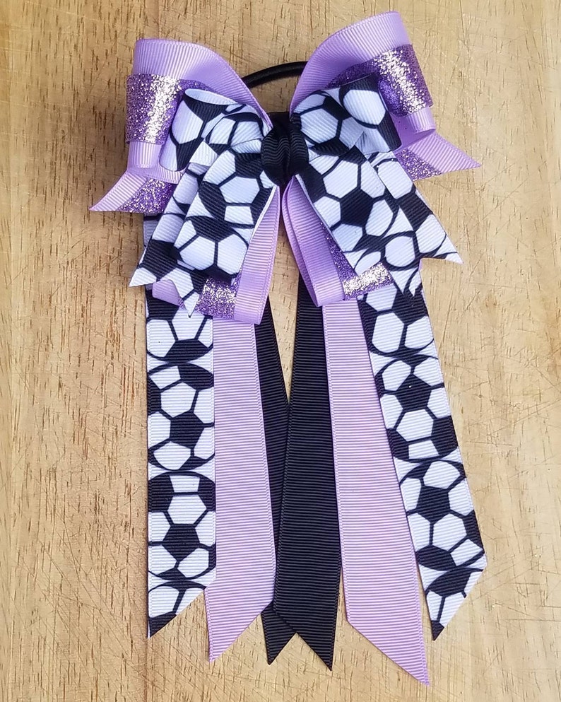 May include: Purple and black hair bow with a soccer ball pattern. The bow has a glitter accent and is attached to a black elastic.