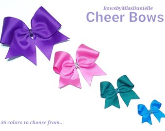 May include: Four satin cheer bows in purple, pink, teal, and blue, each featuring a central rhinestone accent. The text BowsbyMissDanielle Cheer Bows and 36 colours to choose from are displayed in purple font against a plain white background.