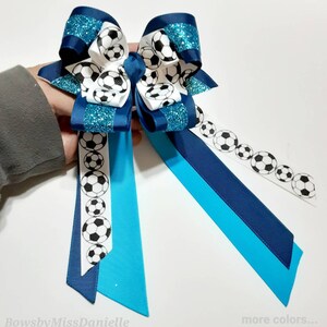 May include: A blue and turquoise hair bow with a white ribbon featuring a soccer ball pattern. The bow has glitter accents.