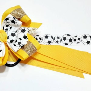 May include: A yellow and white hair bow with a soccer ball pattern. The bow has gold glitter accents and is attached to a black elastic hair tie. The bow is also accompanied by a yellow ribbon with a soccer ball pattern.